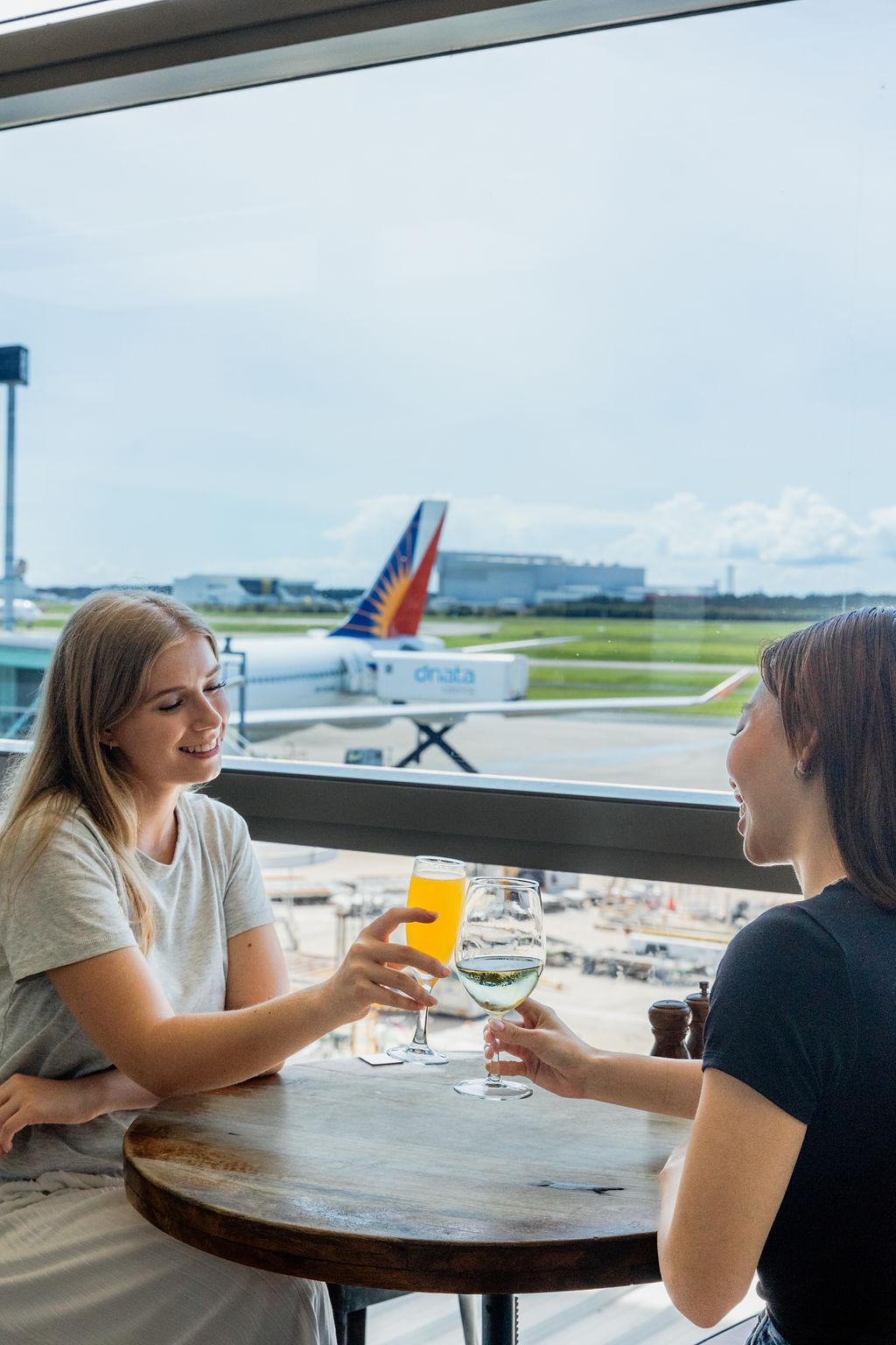 Two women clinking glasses while sitting by the window at the airport