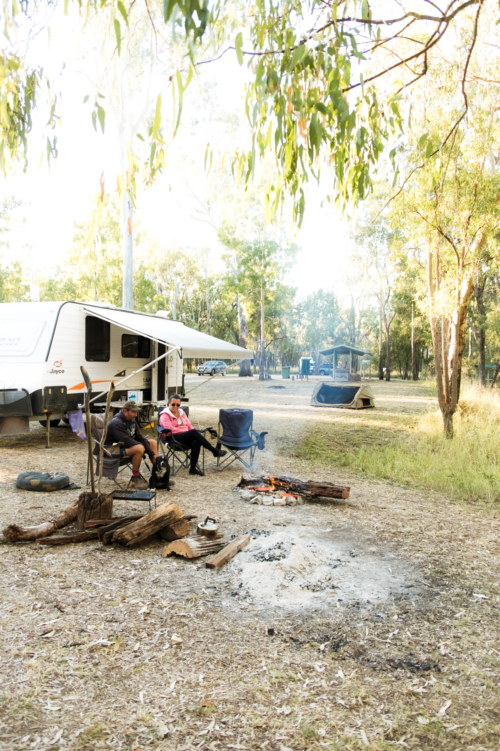 Two people sitting near campfire with camper in background